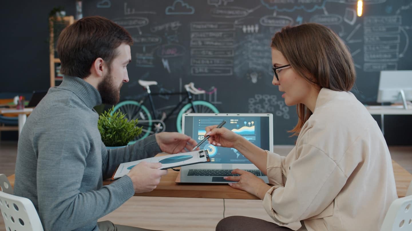 Two business people discuss data on a laptop and document.